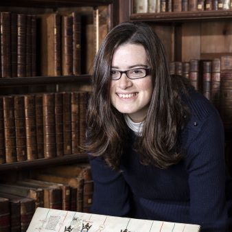 Joanna Bellis headshot in front of bookshelves holding a manuscript
